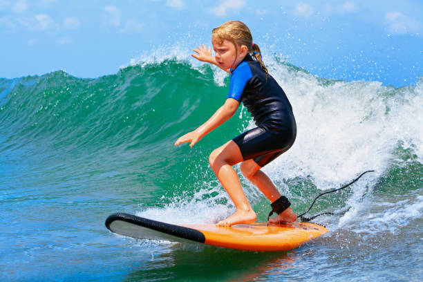 Happy baby girl - young surfer ride on surfboard with fun on sea waves. Active family lifestyle, kids outdoor water sport lessons and swimming activity in surf camp. Summer vacation with child.